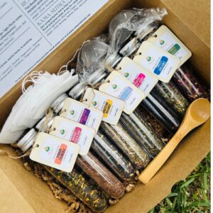 An elegant Tea Sampler Gift Box displayed on a wooden table, featuring a variety of neatly arranged herbal tea blends in individual packaging. The box includes labels showcasing flavors like chamomile, mint, and turmeric ginger, with a warm and inviting aesthetic. A teacup filled with steaming tea is placed beside the box, surrounded by loose tea leaves and a sprig of fresh herbs, evoking a sense of relaxation and wellness.